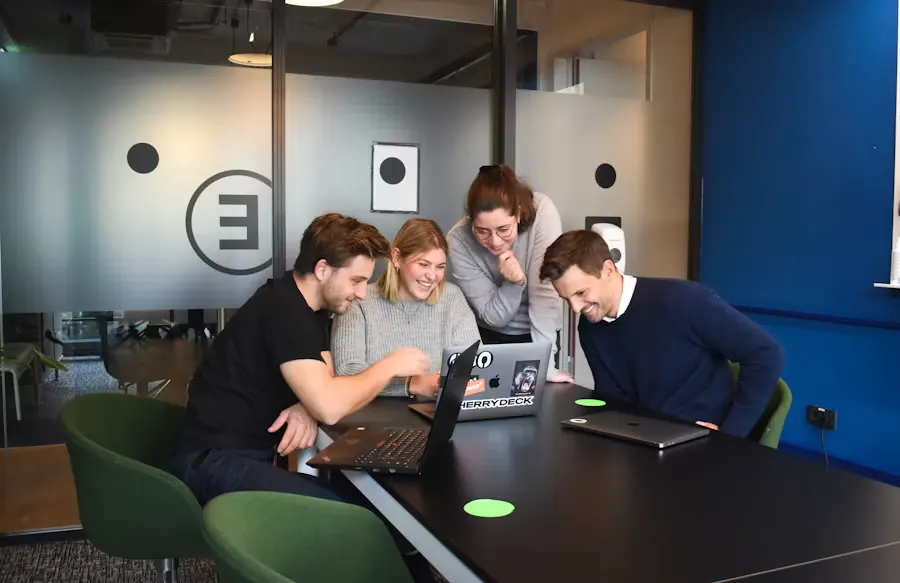 Four colleagues laughing together while looking at a laptop in a meeting room.